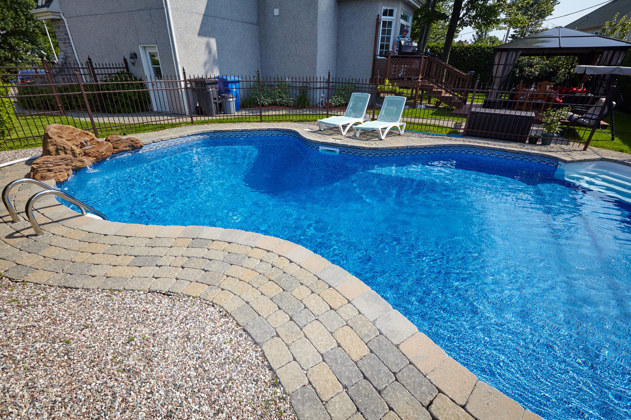 A backyard with a kidney-shaped swimming pool, two white lounge chairs, a shaded seating area, and surrounding greenery.